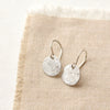 The silver raw silk disc earrings styled on tan linen